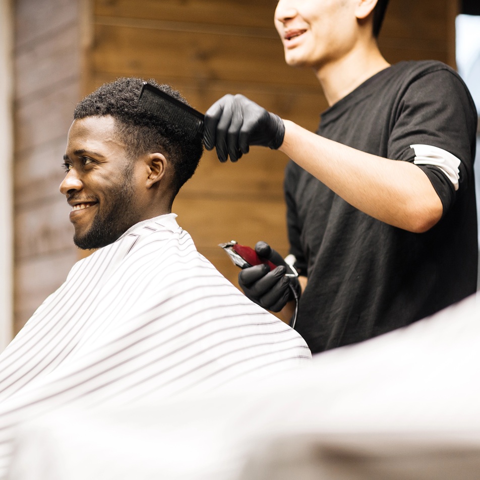 Barber stands behind his client in the barber chair, combing his hair. Both men are smiling.