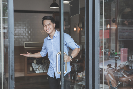Young man holding the door open with one hand, gesturing to come in to his shop with the other.