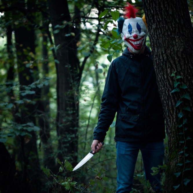 A man in a creepy clown mask is standing partially hidden behind a tree in the woods, holding a big knife.